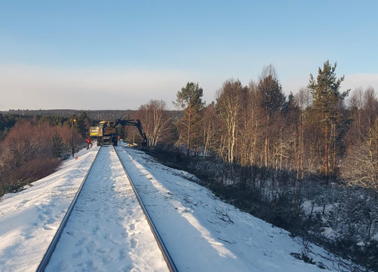 en gravemaskin står på toglinja og driver trefelling langs en jernnbane om vinteren