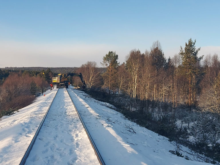 en gravemaskin står på toglinja og driver trefelling langs en jernnbane om vinteren